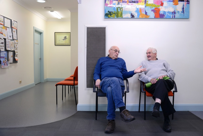 Two elderly men sitting on chairs in a doctor's waiting room, talking