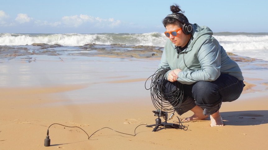 A woman crouches on the sand wearing headphones.
