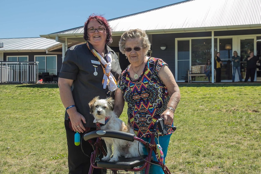 A dog on an aged care walker with a nursing home staff and resident standing behind