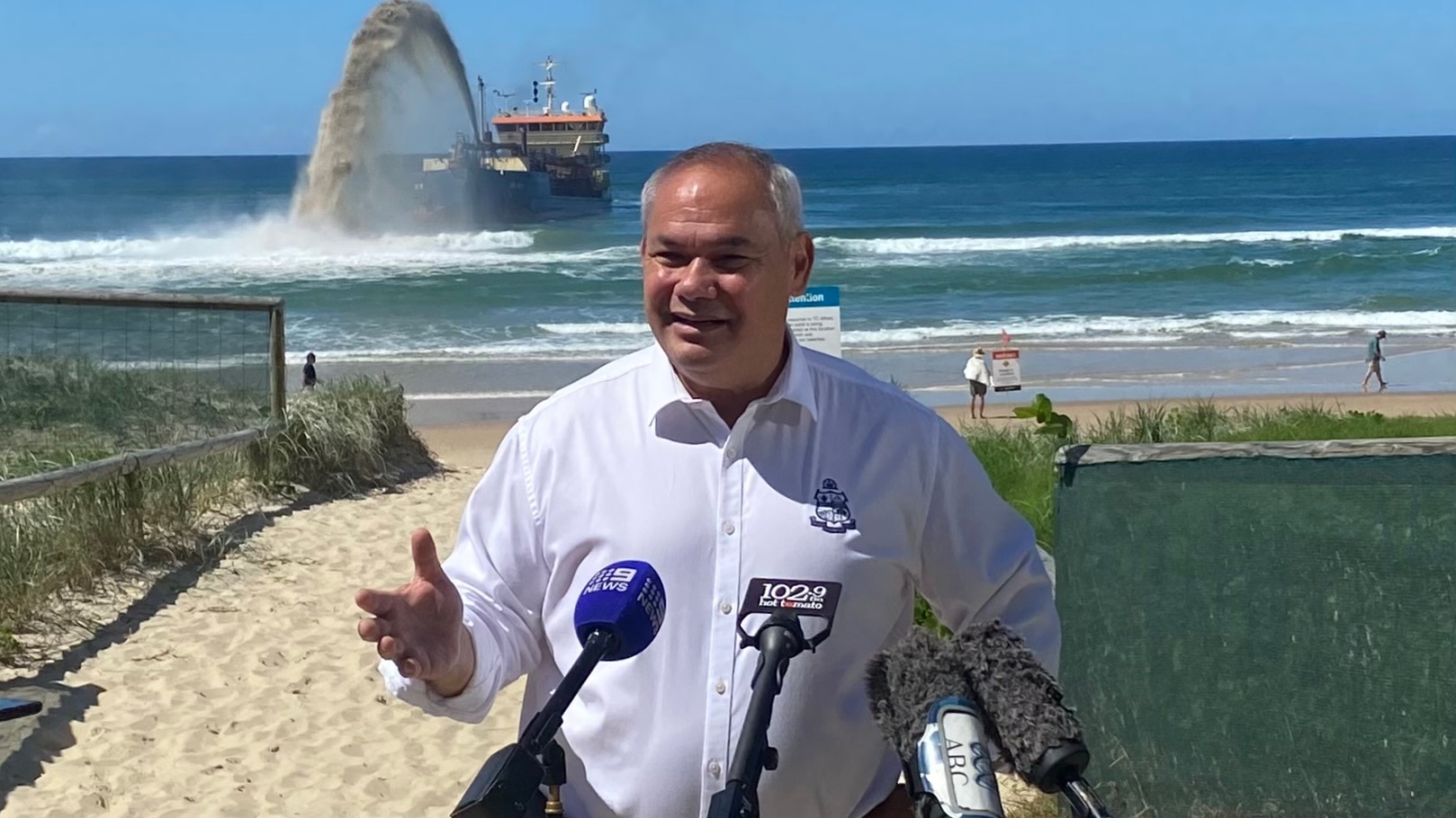 A bald man in a business shirt stands on a beach and speaks to the media.