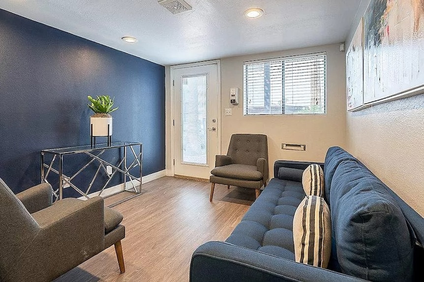 A loungeroom with a long blue couch and painted blue wall. A glass door and window is on the far wall. 