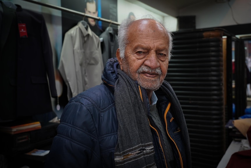 An older man wearing a winter jacket stands inside a tailor's shop