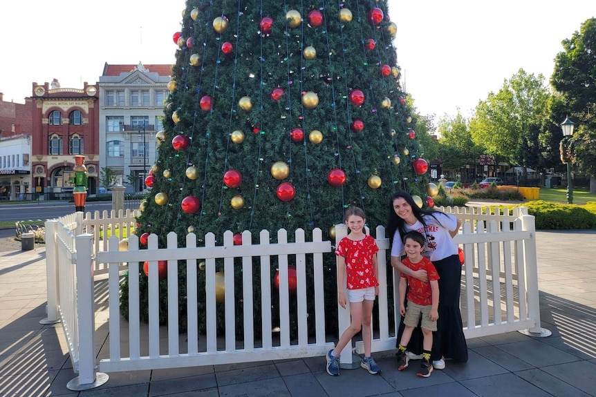 a woman and two young children stand in front of a huge Christmas tree.