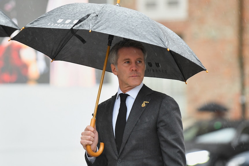 A man dressed in a dark suit holds an umbrella while looking over his shoulder.