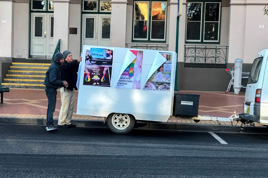 Two men stand at the back of a trailer which displays anti same sex marriage posters.