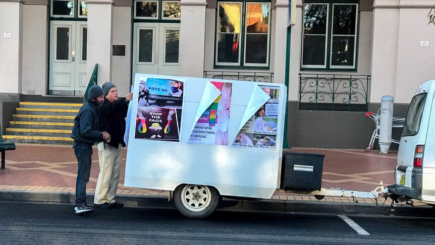 Two men stand at the back of a trailer which displays anti same sex marriage posters