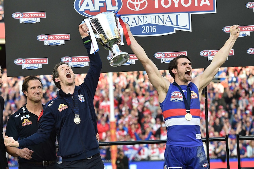 A coach watches as his team's acting captain and injured captain smile as they hold the AFL trophy.