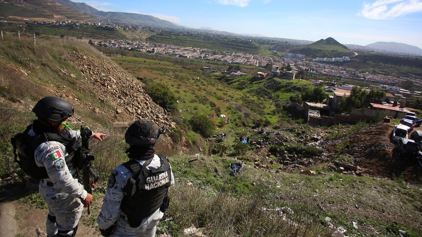 Two armed men in camouflage gear, bulletproof vests and helmets look over a valley.