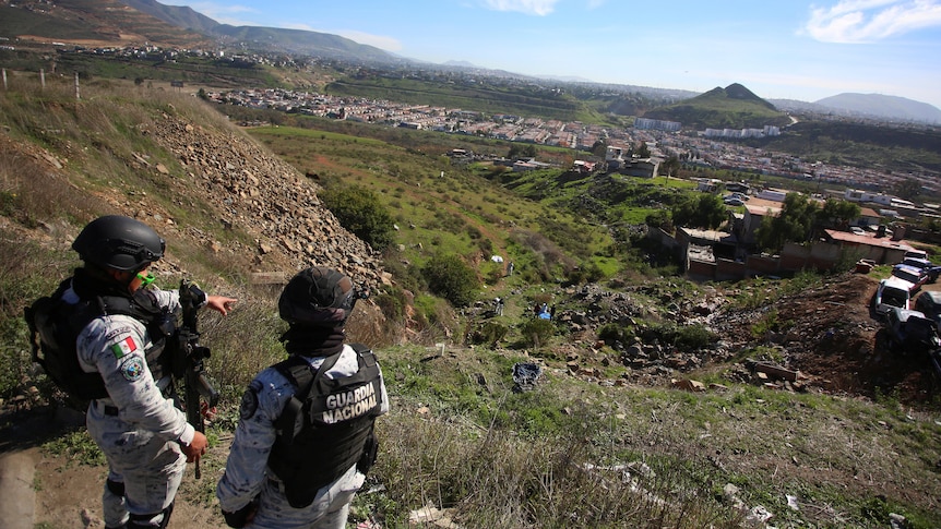 Two armed men in camouflage gear, bulletproof vests and helmets look over a valley.