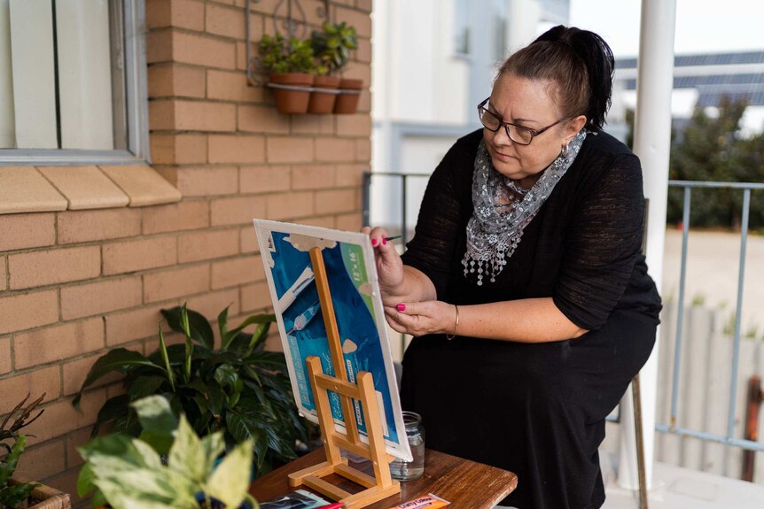 A woman seated painting on a small easel on an apartment balcony.