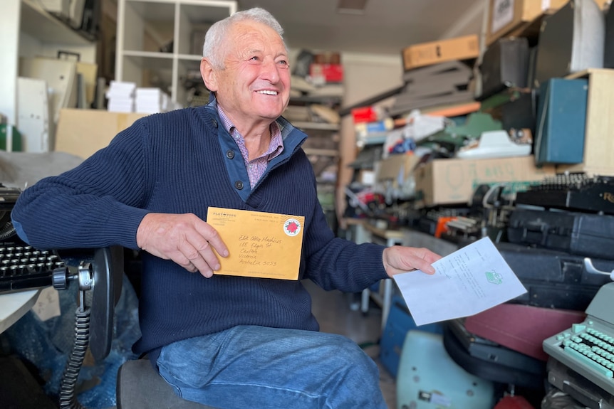 An elderly man holds a letter in one hand and an envelope in the other while sitting in his typewriter repair workshop.