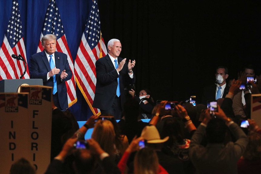 Two men stand on stage wearing suits and clapping at a crowd gathered in front of them.