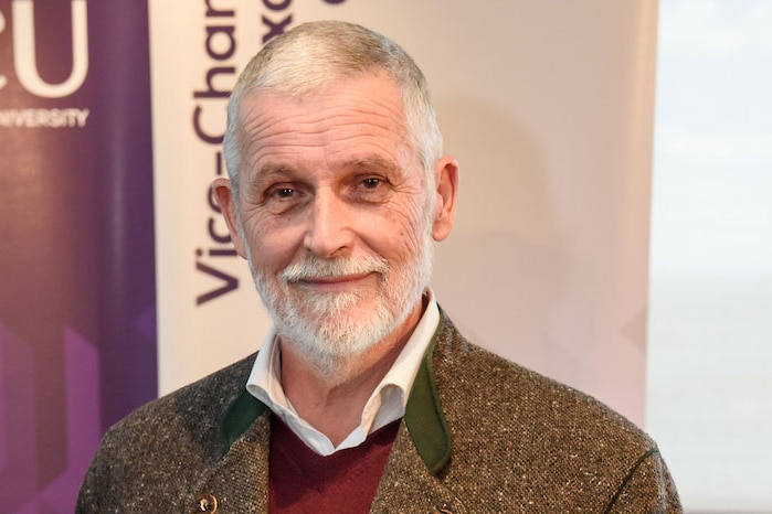 A man with grey hair and a beard smiles in front of purple and white banners.