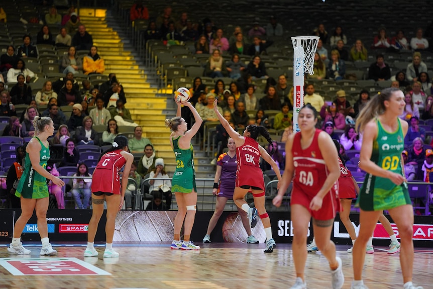 An Australian netball player is lifting a netball above her head to try to score, a Tongan player defends.