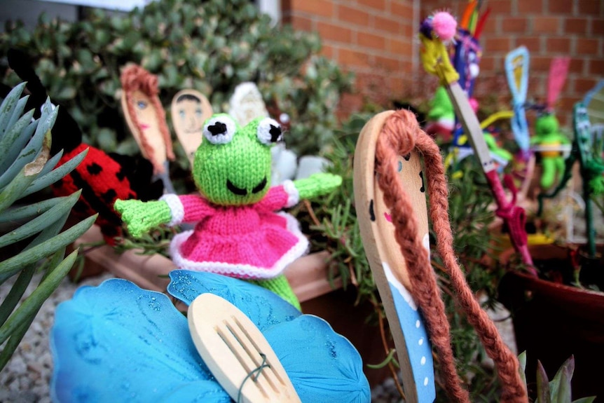 A knitted green frog decorations and smaller wooden crafts of people stick up between plants in a garden.