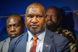 A close up of James Marape wearing a red tie and dark blue suit at a podium infront of a crowd.