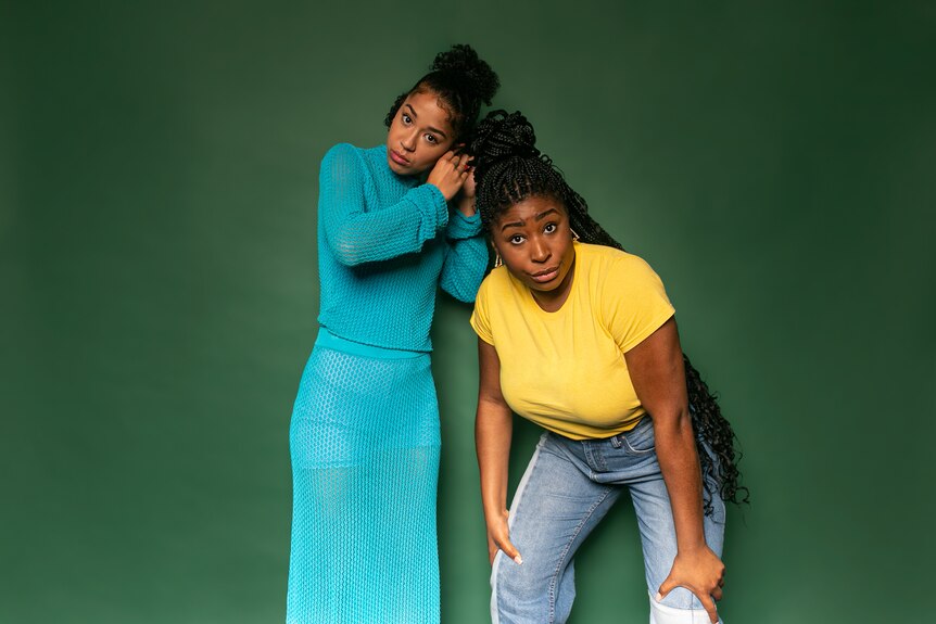 Two Black women stand in front of a green background, one adjusting her earring, the other bent forward. 