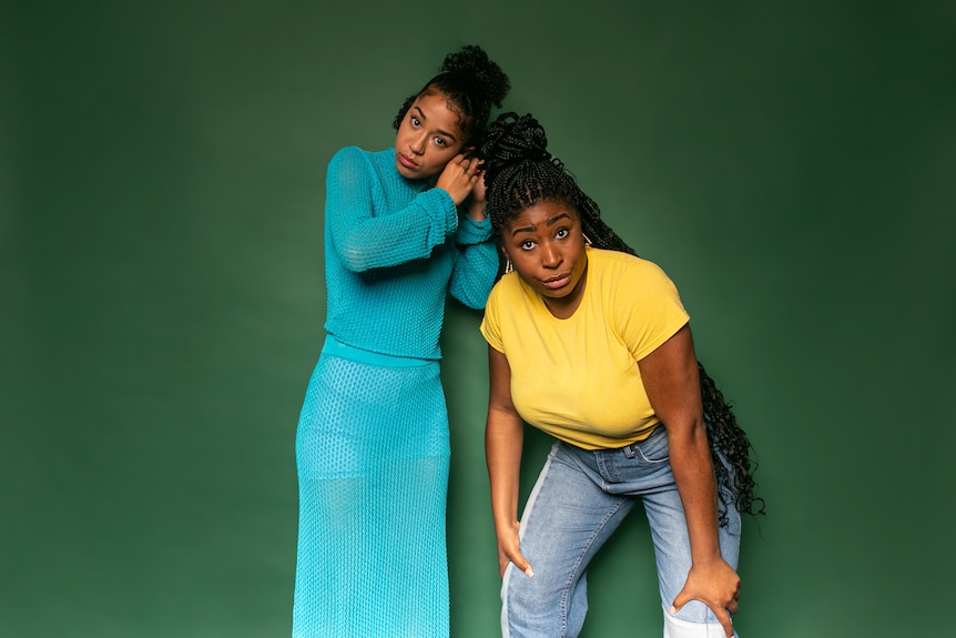 Two Black women stand in front of a green background, one adjusting her earring, the other bent forward. 