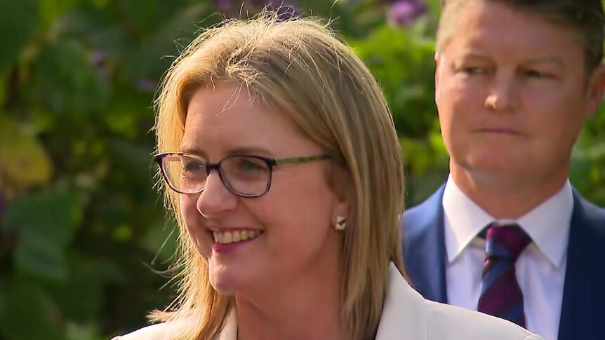 Jacinta Allan wears a white jacket and glasses and smiles while Ben Carroll stands behind her with a tight lipped smile.