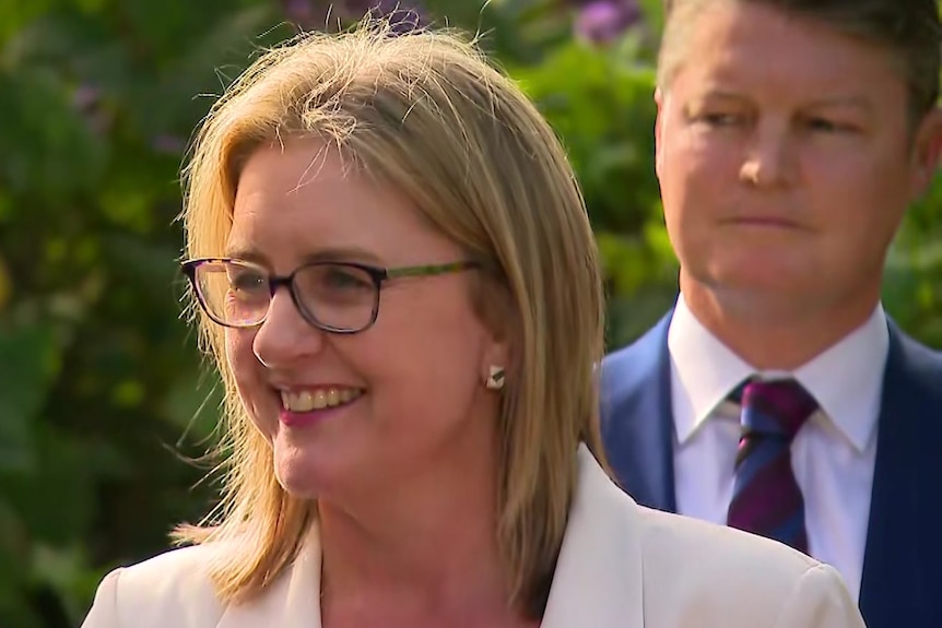 Jacinta Allan wears a white jacket and glasses and smiles while Ben Carroll stands behind her with a tight lipped smile.