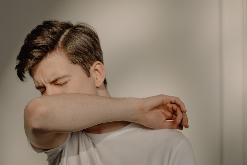 A close-up photo of a man covering his face with his arm while coughing