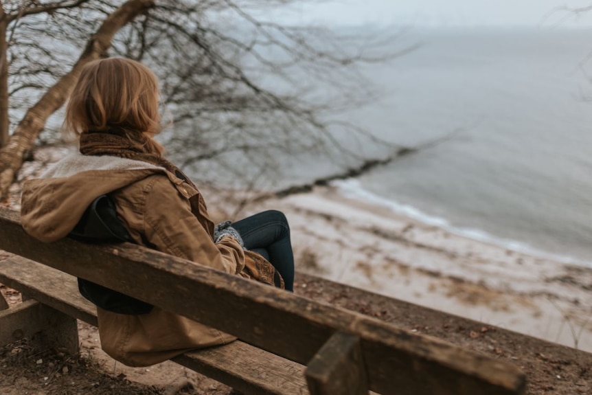 The back of a woman wearing warm jacket, scarf and jeans, sitting on a wooden bench facing beach and sea below.