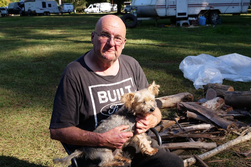 Man sits in a camping chair with his small, cream-coloured dog on his lap, firewood and caravan in the background.