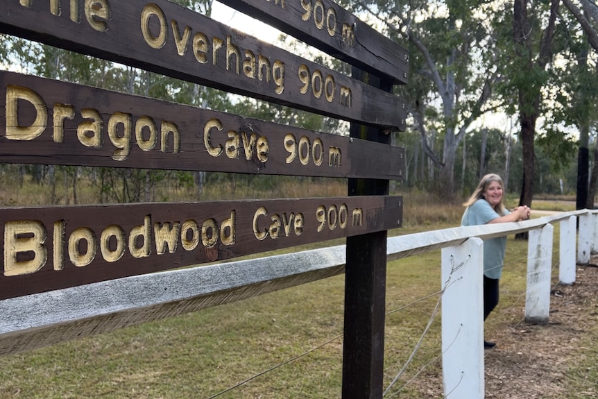 A wooden sign detailing walking trails in the foreground, with a woman leaning against a fence in the background,
