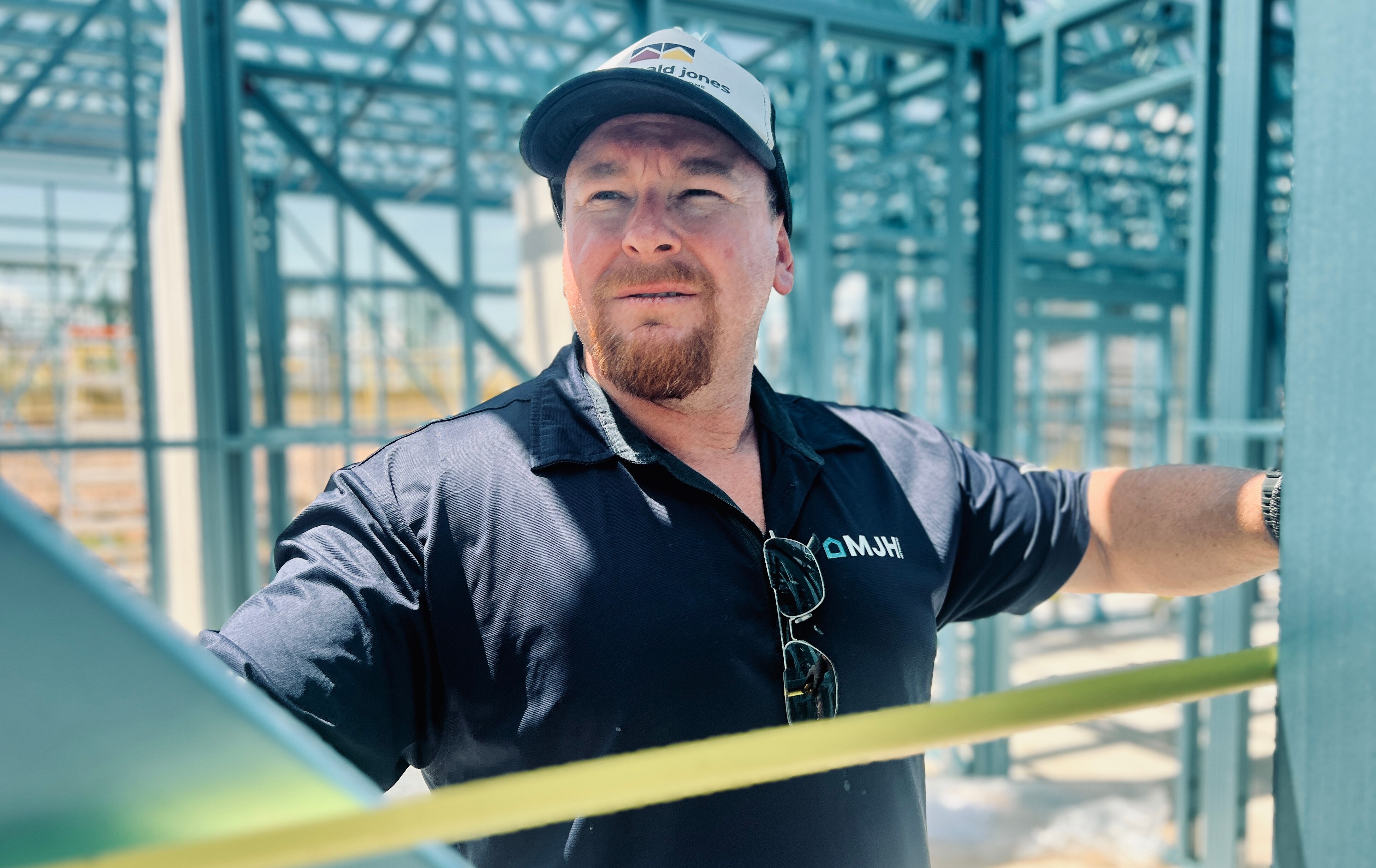 Serious Brian looks ahead, wears cap, blue polo tee with MJH logo, stands inside a house frame being built.