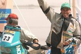 a motorcyclist raises arm in victory standing next to motorbike in Dakar rally