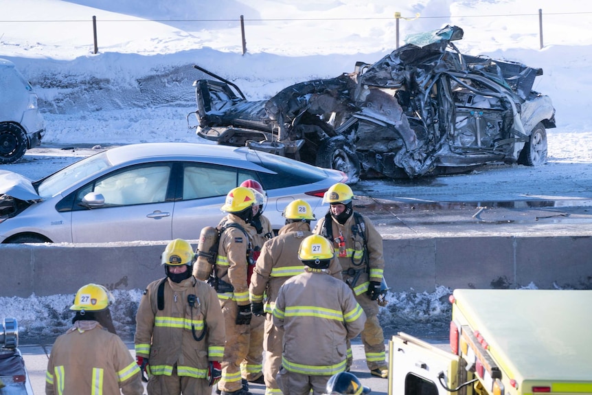Emergency crews look on as a crumpled car's chassis sits on a snowy highway