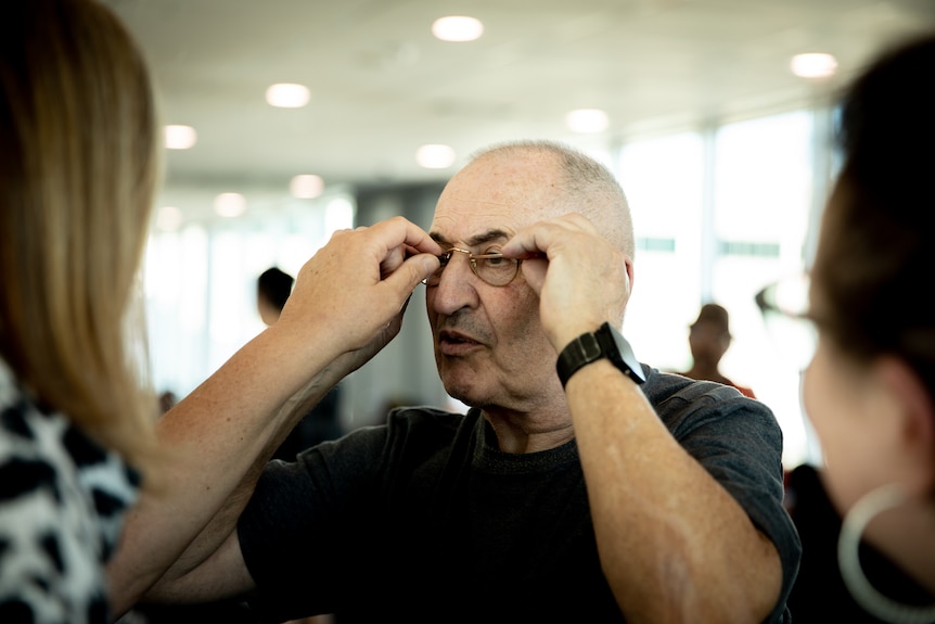 An older man tries on a pair of gold optical glasses, with a woman assisting him, inside an airport.