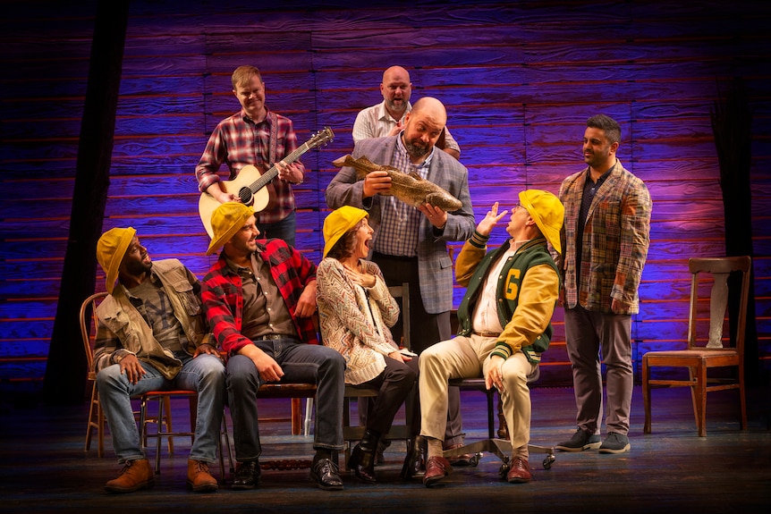 Musical cast members on stage, four of them with yellow hard hats sitting in chairs, one of them plays a guitar.