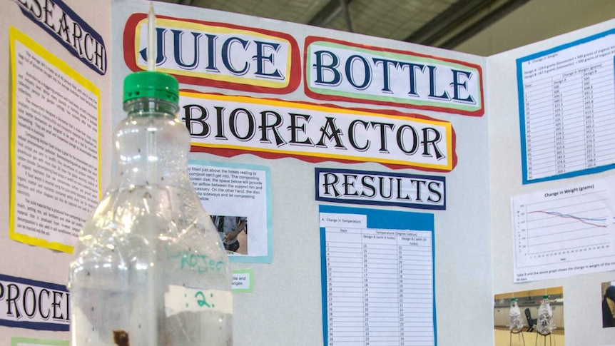 A plastic bottle on a stage with a large explanation written on boards behind it.