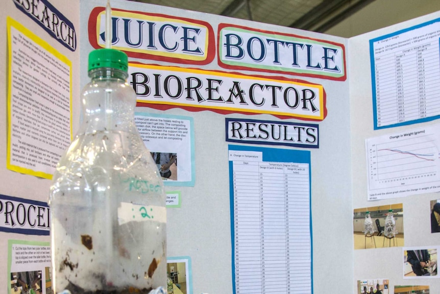 A plastic bottle on a stage with a large explanation written on boards behind it.