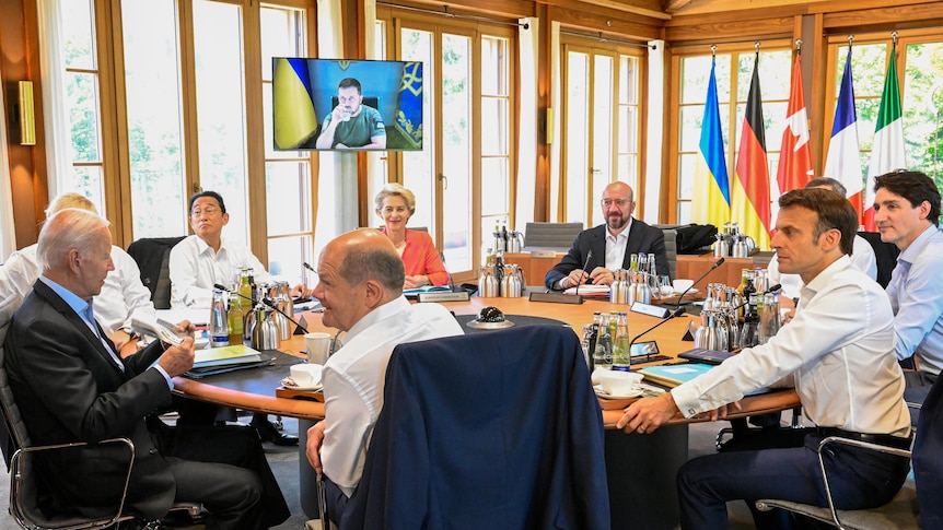 G7 leaders sitting around a table with the Ukrainian president on a monitor in the background.