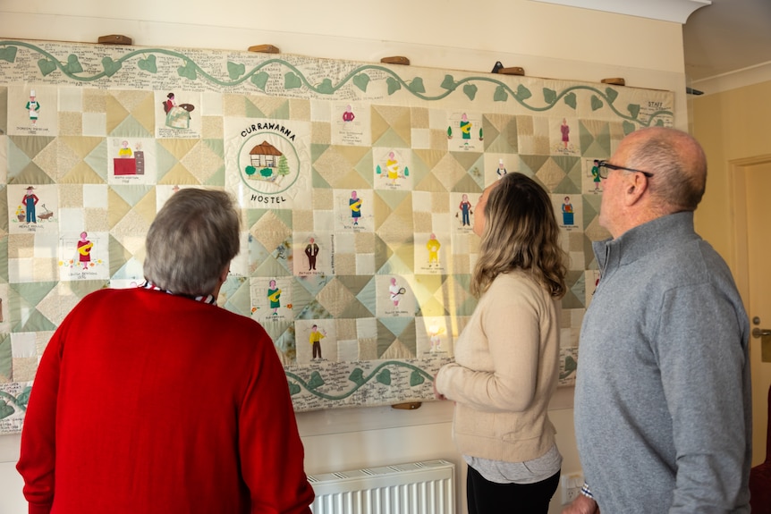 Three people looking at a quilt hung up on a wall. 