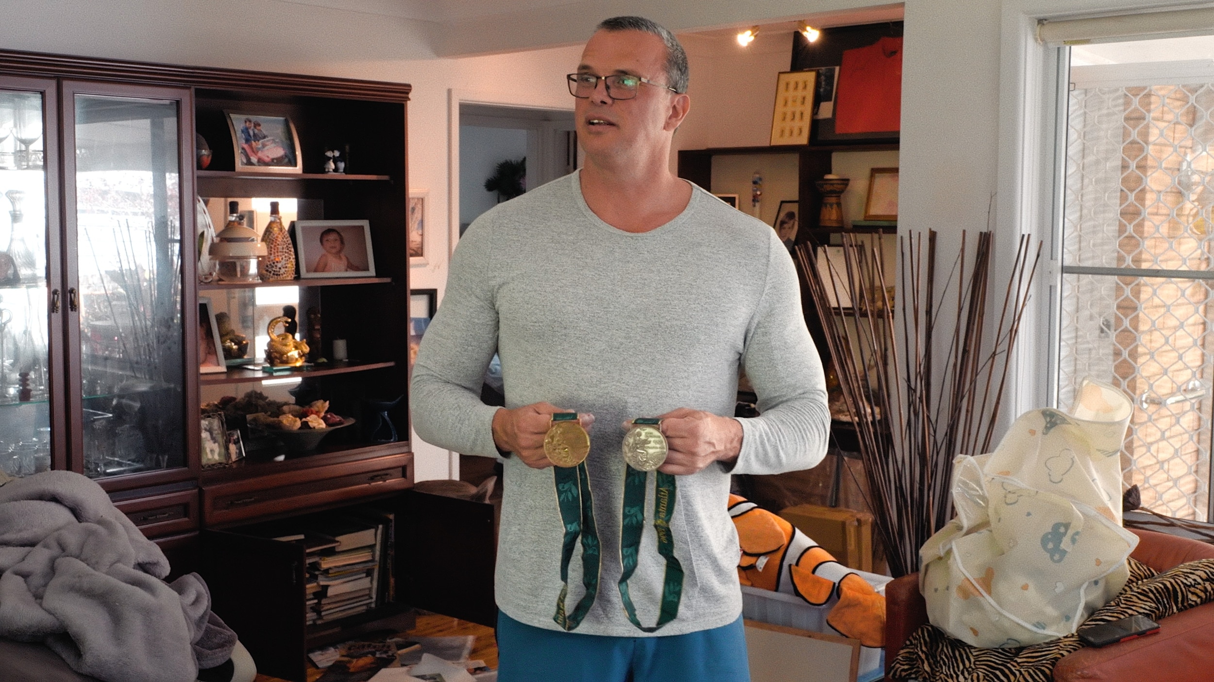 A man wearing a long sleeved grey shirt and glasses holds a silver and bronze medal standing in his living room.