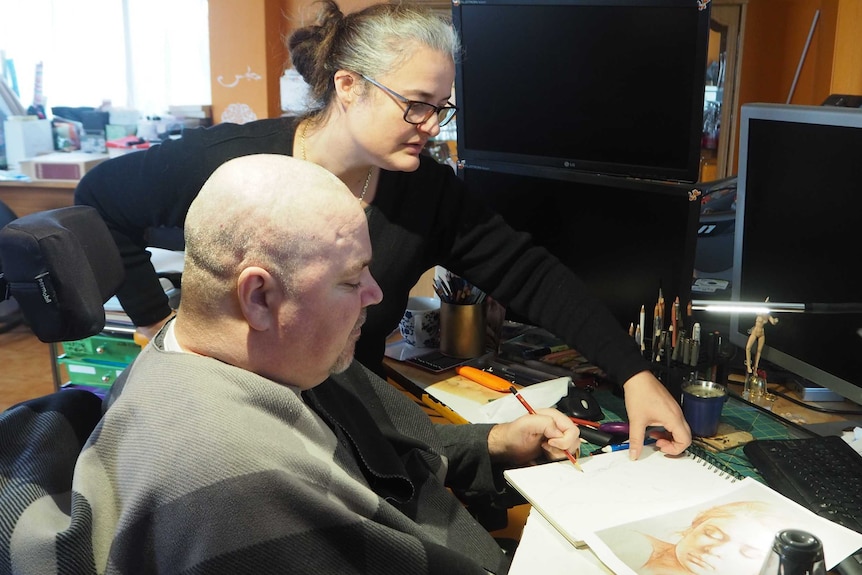 A woman helps a man with tetraplegia with a drawing.