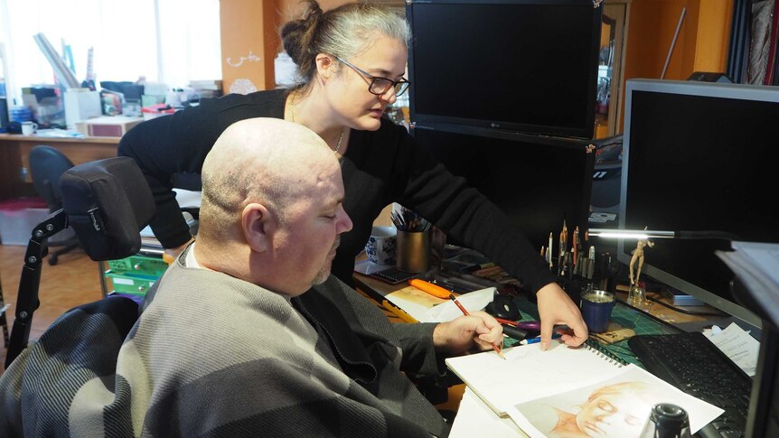 A woman helps a man with tetraplegia with a drawing.