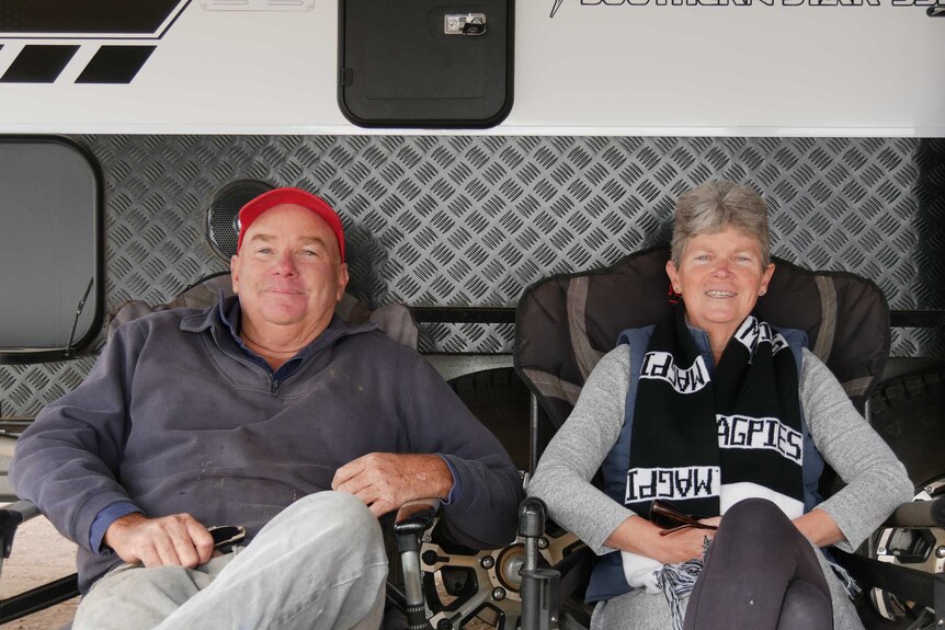 An older couple sit in camping chairs in front of the side of a caravan, smiling for the camera.