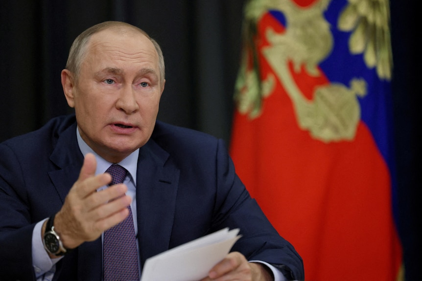 Russian President Vladimir Putin holds papers while speaking in front of Russian flag.