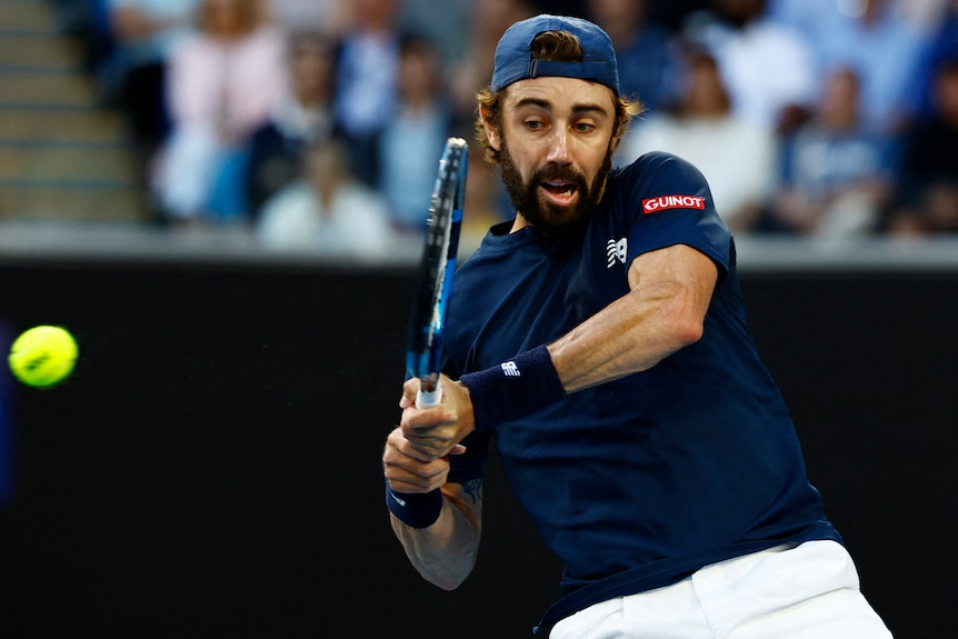 留着胡子的男子网球运动员，穿着深色衬衫，在一场夜间比赛中，双手反手击球。