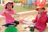 Two girls in pink play in a sandpit with pots and pans and toys.