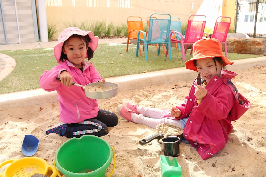 Two girls in pink play in a sandpit with pots and pans and toys.
