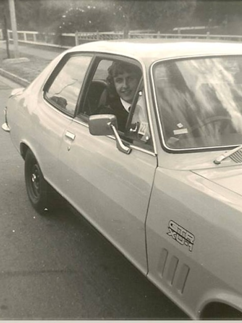 A black and white photograph of a man driving a car, he can be seen smiling through the driver's side window