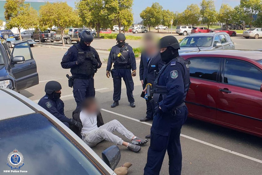 Five police officers, four masked and one with blurred face, surround man wearing white clothes sitting on ground, face blurred