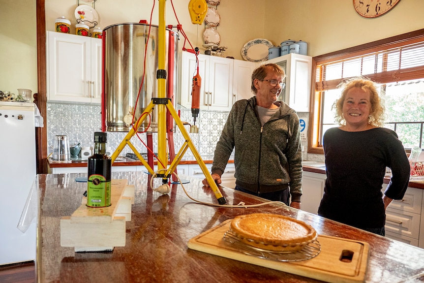 A man and woman stand in a kitchen with a pie in front of them.