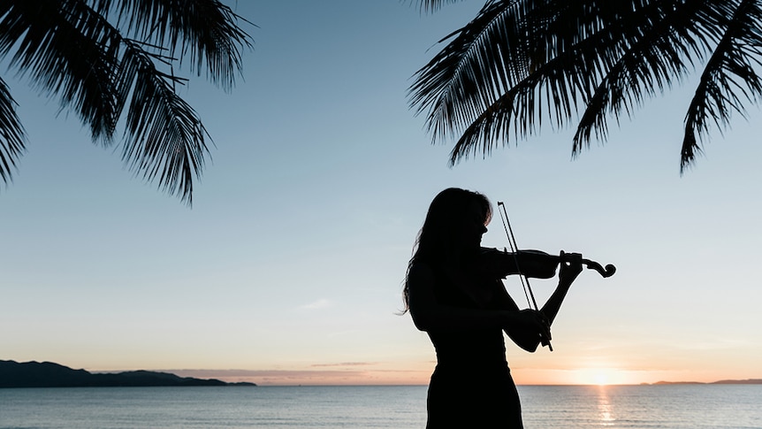The silhouette of a woman playing violin at dusk with palm trees and the sun setting over water in the background.