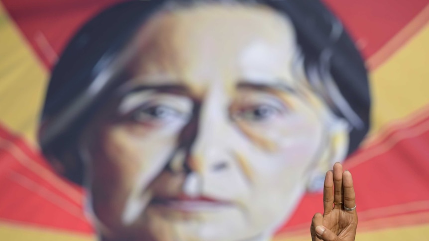 A person shows the three-fingers salute in front of a placard with the image of Aung San Suu Kyi.